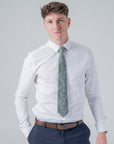 Man wearing a white shirt and navy trousers with a deep green floral patterned cotton tie in a studio setting
