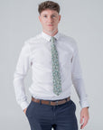 Man wearing Elmwood Bloom cotton tie with forest green floral pattern, white shirt, and navy trousers against plain background
