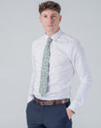 Man wearing Elmwood Bloom cotton tie with green floral pattern, white shirt, navy pants, and brown belt against plain background
