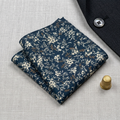 A folded navy blue pocket square with a delicate white and beige floral pattern, styled on a textured light gray linen surface next to a dark suit jacket and a gold thimble.